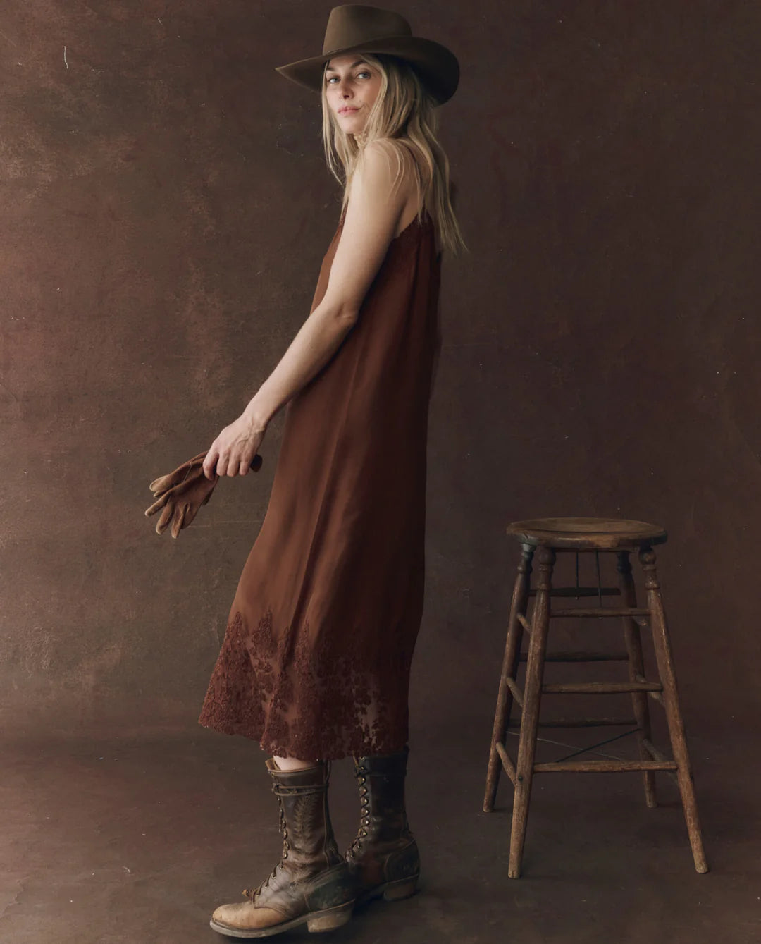 A woman in The Great Inc.'s Legacy Dress, a vintage-inspired brown sleeveless design, stands by a wooden stool holding brown gloves, styled with a brown hat and lace-up boots against a brown textured backdrop.