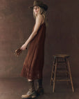 A woman in The Great Inc.'s Legacy Dress, a vintage-inspired brown sleeveless design, stands by a wooden stool holding brown gloves, styled with a brown hat and lace-up boots against a brown textured backdrop.