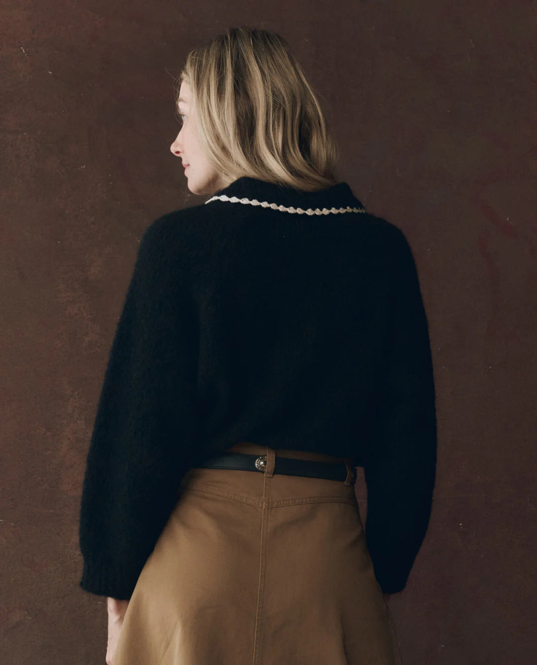 A woman with blonde hair wears The Great Inc.&#39;s The Crochet Collar Pullover, a black sweater with a crochet collar, and a tan skirt as she stands facing away from the camera against a plain brown background.