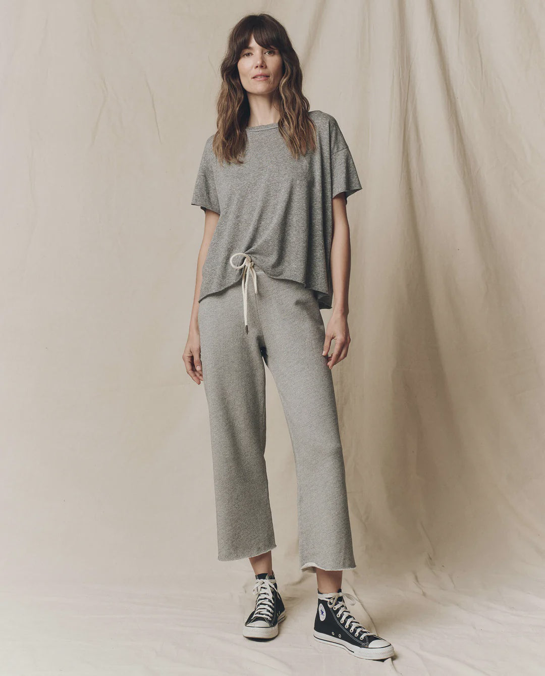 A woman with wavy brown hair stands against a beige backdrop, wearing The Great Inc. Cropped Sweat Pant in grey cotton terry with a matching t-shirt and black Converse high-tops, looking relaxed.