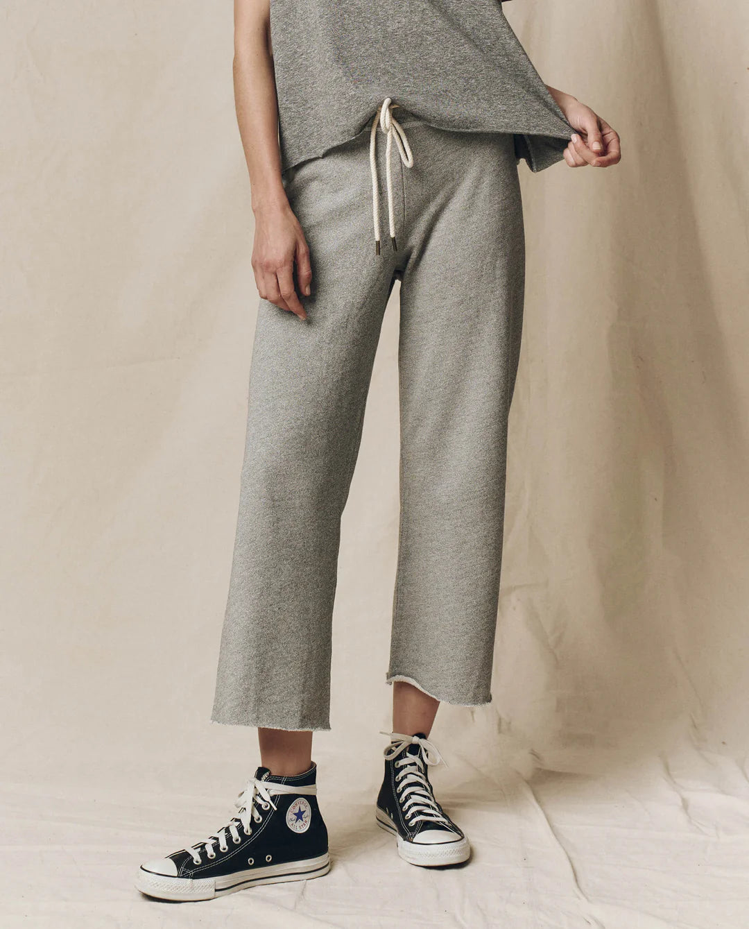A person models The Great Inc. Cropped Sweat Pant in grey washed cotton terry with a matching top and black high-top Converse, standing against a neutral background and lifting one side of their shirt. Their face is not visible.