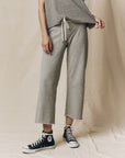 A person models The Great Inc. Cropped Sweat Pant in grey washed cotton terry with a matching top and black high-top Converse, standing against a neutral background and lifting one side of their shirt. Their face is not visible.