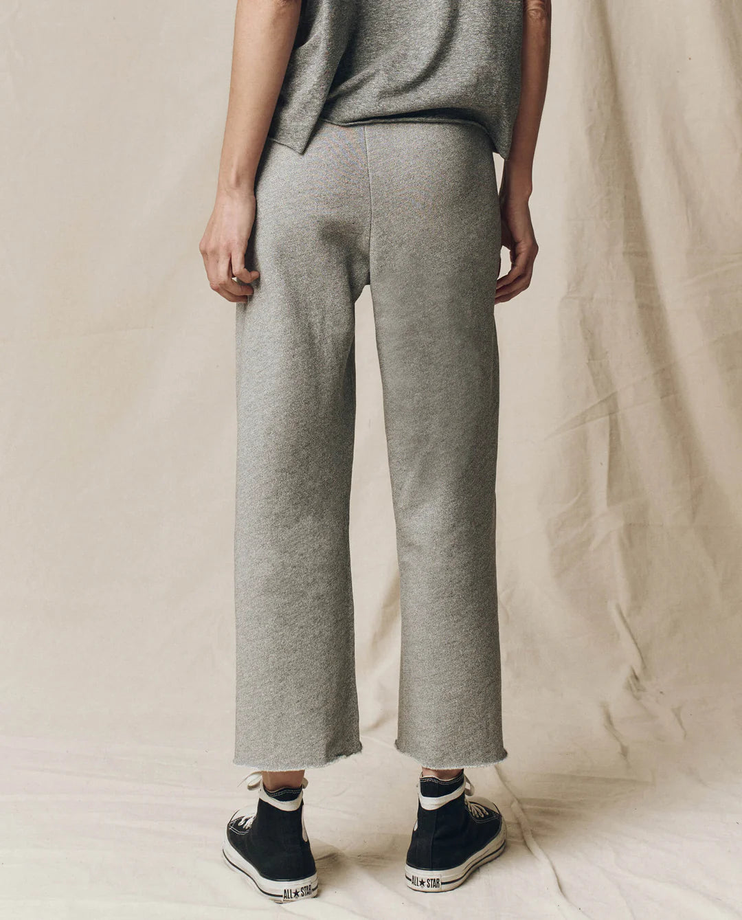 A person stands facing away, wearing The Great Inc. Cropped Sweat Pant in grey with a drawstring waist and raw hems, paired with a matching short-sleeve top and black high-top sneakers against a light beige fabric background.