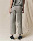 A person stands facing away, wearing The Great Inc. Cropped Sweat Pant in grey with a drawstring waist and raw hems, paired with a matching short-sleeve top and black high-top sneakers against a light beige fabric background.