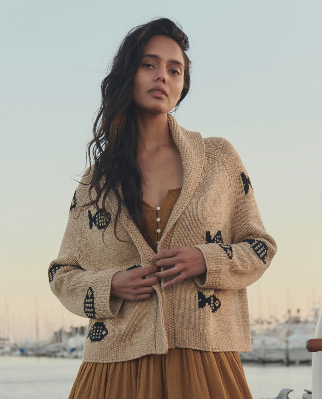 A woman with long wavy hair wears The Great Inc.’s "The Swimming Fish Lodge Cardigan," featuring a black swimming fish motif in beige, over a brown dress. She stands outdoors by a marina at sunset, boats visible in the background.