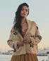 A woman with long wavy hair wears The Great Inc.’s "The Swimming Fish Lodge Cardigan," featuring a black swimming fish motif in beige, over a brown dress. She stands outdoors by a marina at sunset, boats visible in the background.