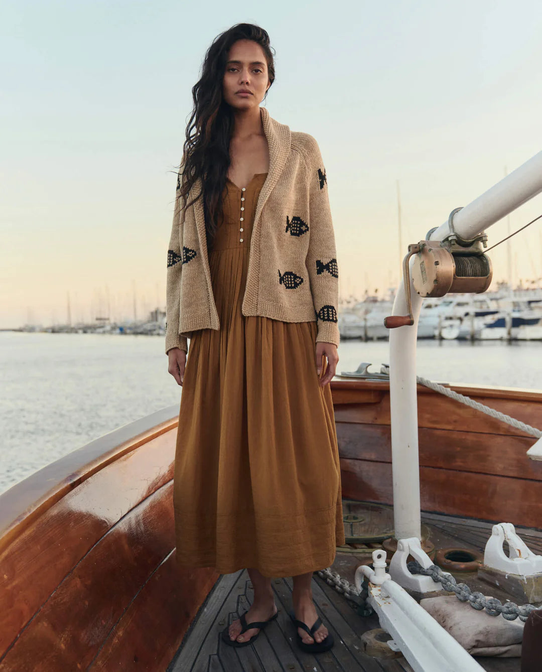 A woman stands on a wooden boat wearing The Swimming Fish Lodge Cardigan by The Great Inc. over a long brown dress and black sandals, with sailboats filling the sunset-lit marina in the background.
