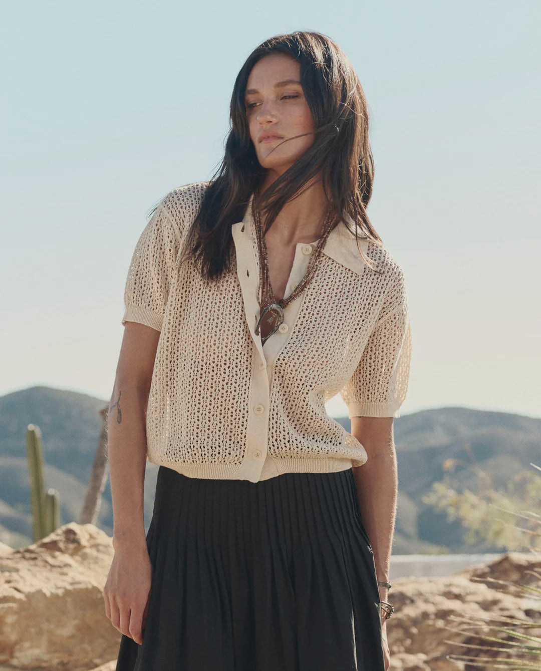 A woman with long dark hair stands outdoors among desert cacti, wearing The Great Inc.'s The Crochet Polo—a cream, hand-crocheted short-sleeve cardigan—over a white top and black pleated skirt.