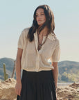 A woman with long dark hair stands outdoors among desert cacti, wearing The Great Inc.'s The Crochet Polo—a cream, hand-crocheted short-sleeve cardigan—over a white top and black pleated skirt.