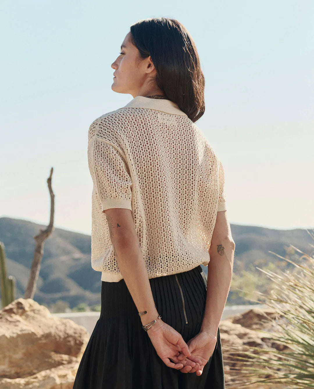 A person with long dark hair stands outdoors, hands clasped behind their back, wearing The Crochet Polo by The Great Inc. in cream, paired with a black skirt, with desert hills and cacti in the background.