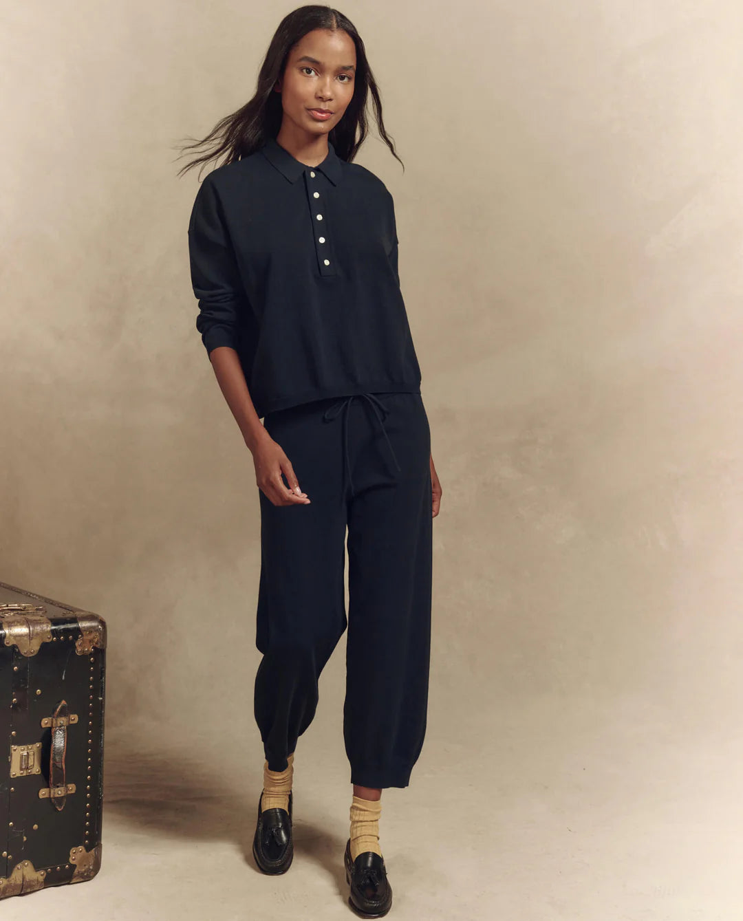 A person stands wearing The Great Inc.'s dark Polo Sweater with matching jogger pants. They sport light-colored socks and black loafers. A vintage trunk on the left showcases timeless menswear against a soft beige background, enhancing the classic style.