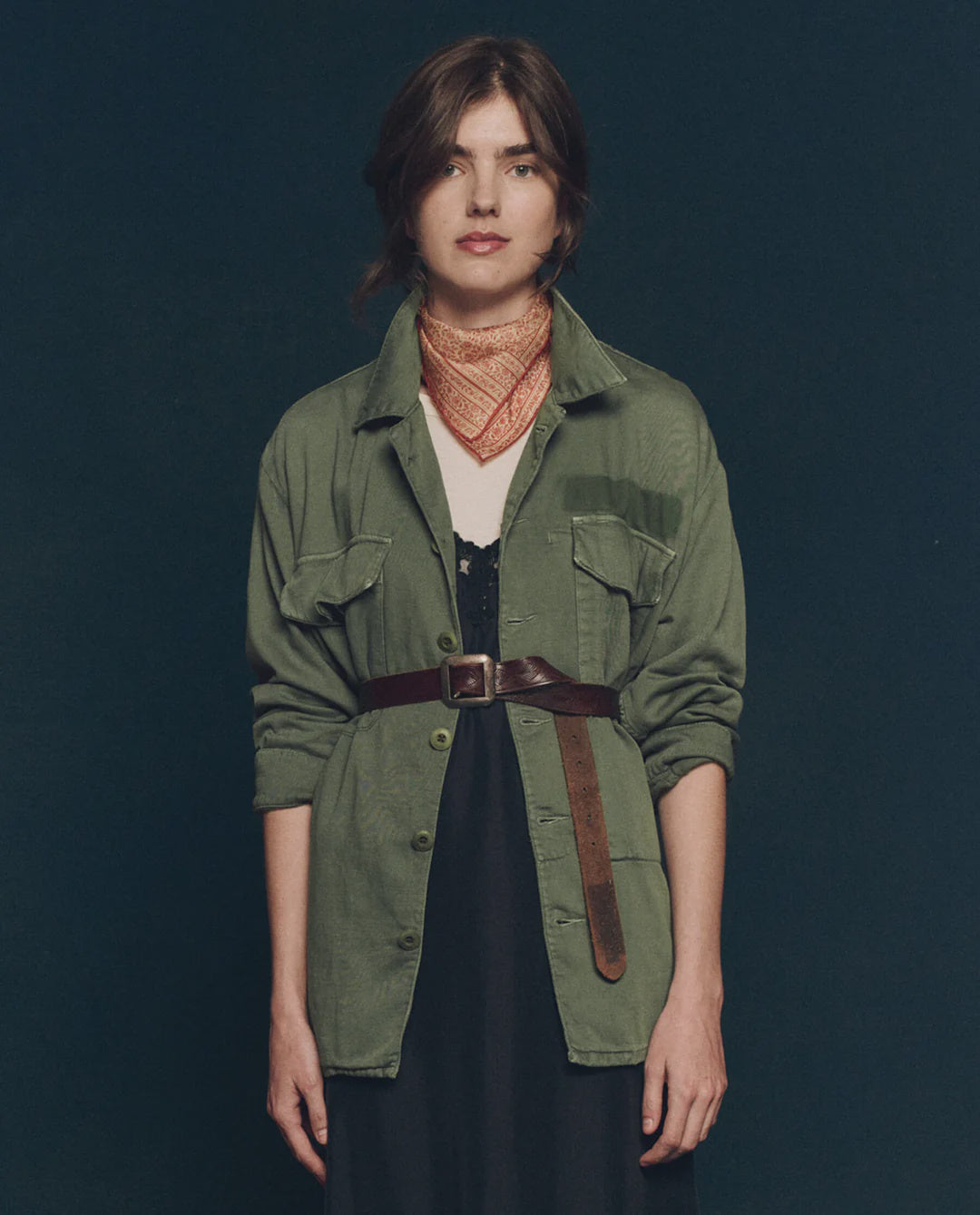 A woman with loosely styled brown hair and a neutral expression stands against a dark background, wearing The Vintage Army Jacket by The Great Inc. in cotton slub twill, belted over a black dress with a patterned red neck scarf.