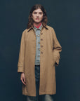 Against a dark backdrop, an individual wears "The Trench Coat" by The Great Inc. A tan cotton canvas piece layered over a denim jacket, creating an A-line silhouette. A red bandana accents their neck as they tuck hands in pockets, focusing intently on the camera.