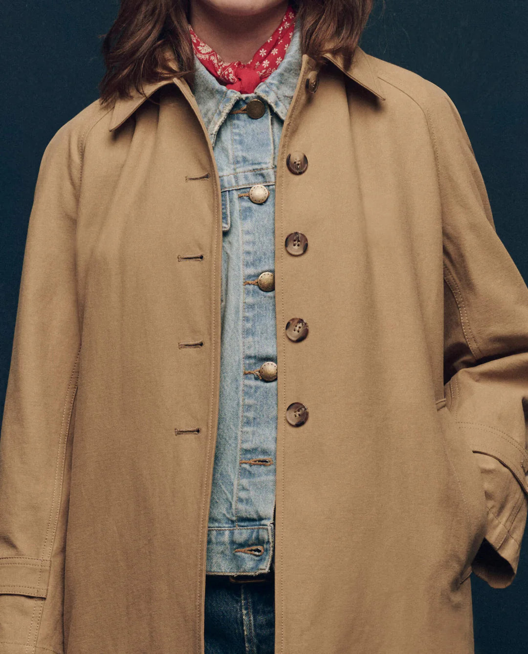 A person wears The Great Inc.'s "The Trench Coat" in beige cotton canvas over a denim jacket with gold buttons, accessorized with a red bandana scarf. The scene features a plain dark background, obscuring the person's face.