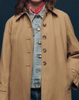 A person wears The Great Inc.'s "The Trench Coat" in beige cotton canvas over a denim jacket with gold buttons, accessorized with a red bandana scarf. The scene features a plain dark background, obscuring the person's face.