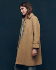 Wearing The Trench Coat by The Great Inc., a person stands against a dark background. Their mid-length brown hair contrasts with the rolled-up cuffs of the cotton canvas trench, revealing denim underneath, as they gaze to the side.