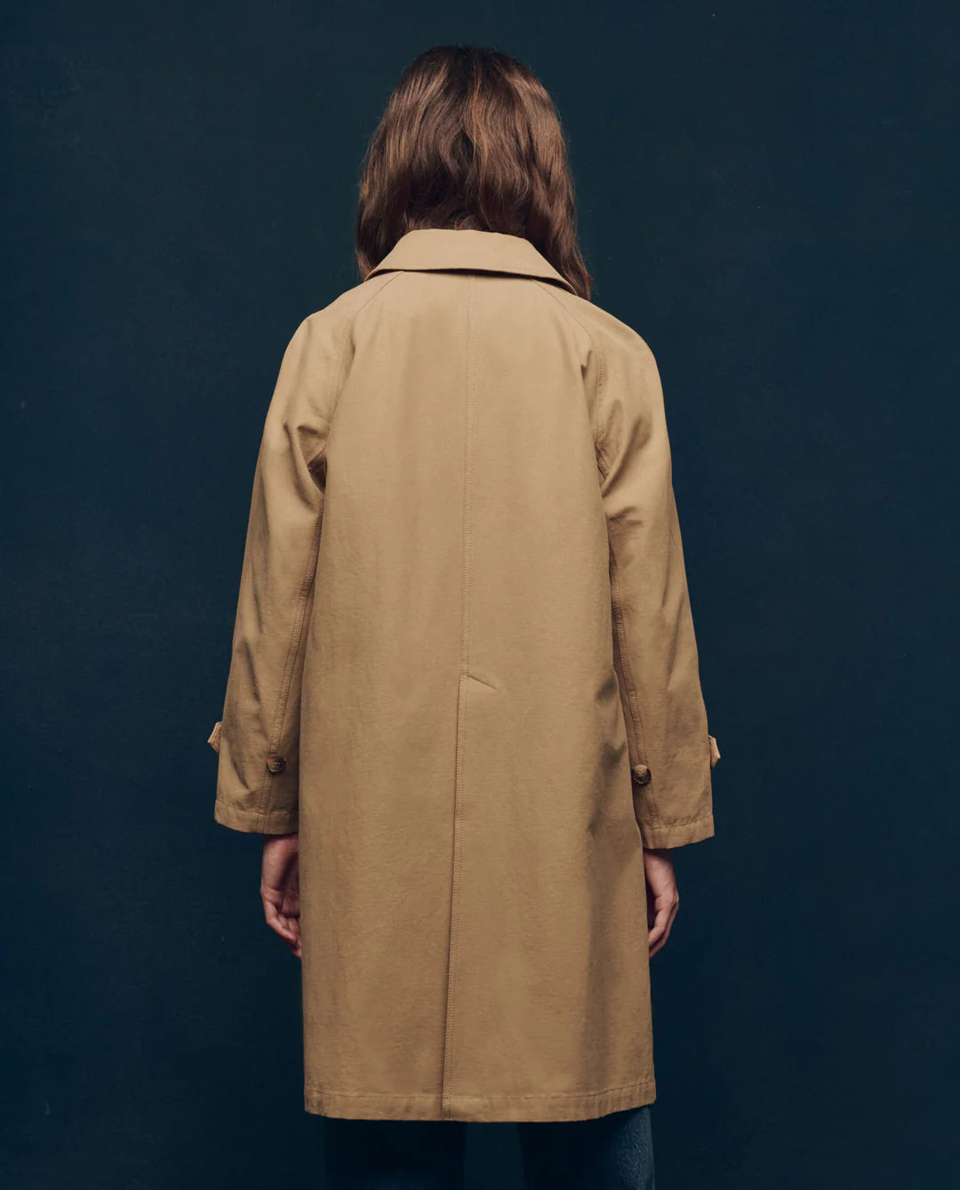 A person with long brown hair, facing away, wears The Trench Coat by The Great Inc., featuring a tan color and A-line silhouette. The background is a plain dark wall.