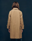 A person with long brown hair, facing away, wears The Trench Coat by The Great Inc., featuring a tan color and A-line silhouette. The background is a plain dark wall.