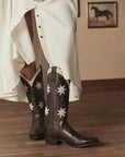 A person wearing a flowing white dress and The Great Inc. Leather Floral Cowboy Boots stands on a wooden floor; a framed horse picture is visible on the wall in the background.
