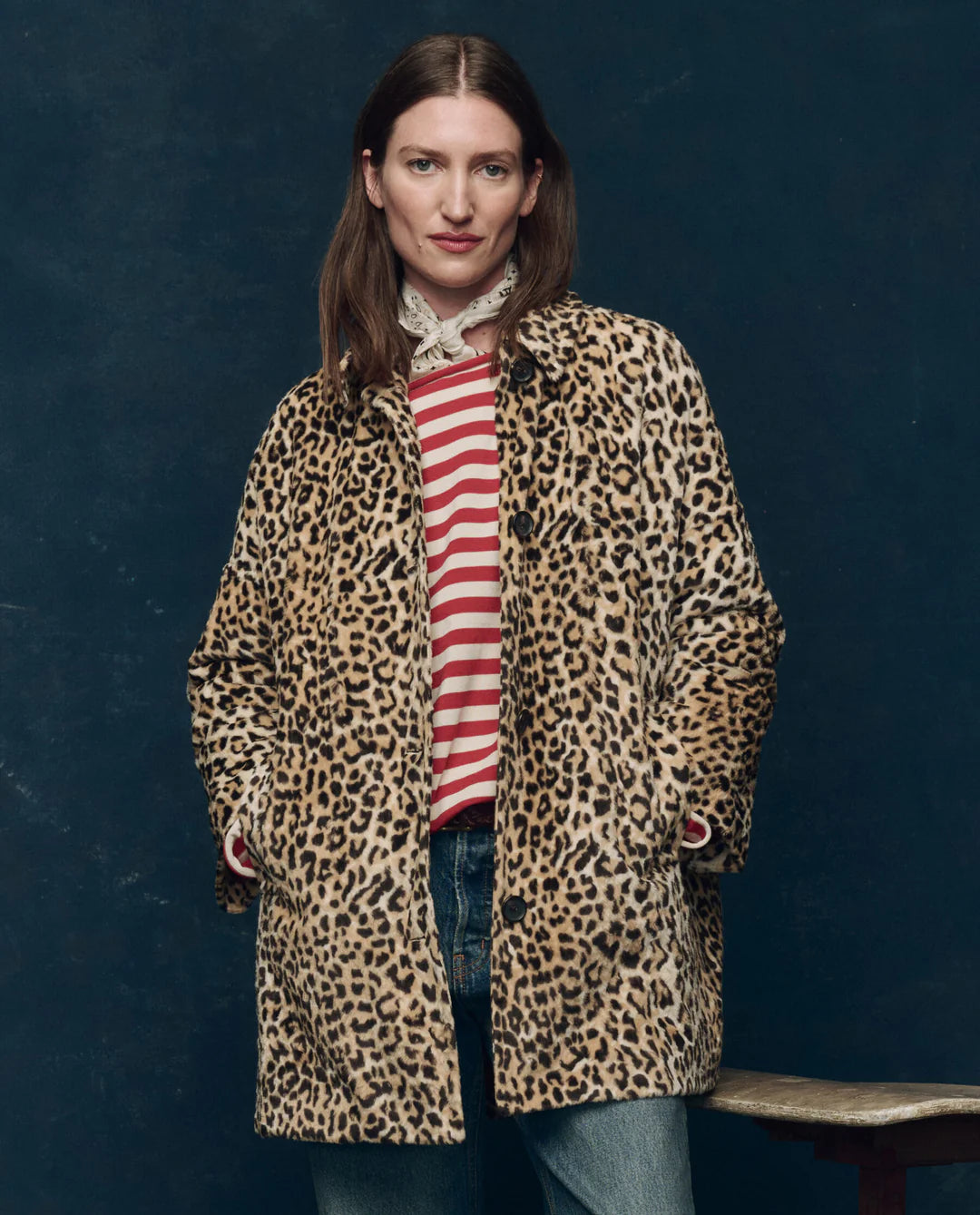 A woman with straight brown hair stands against a dark background, wearing The Car Coat by The Great Inc.—a crushed faux fur leopard print midcentury coat with quilted satin lining—over a striped shirt and blue jeans, hands in her coat pockets.