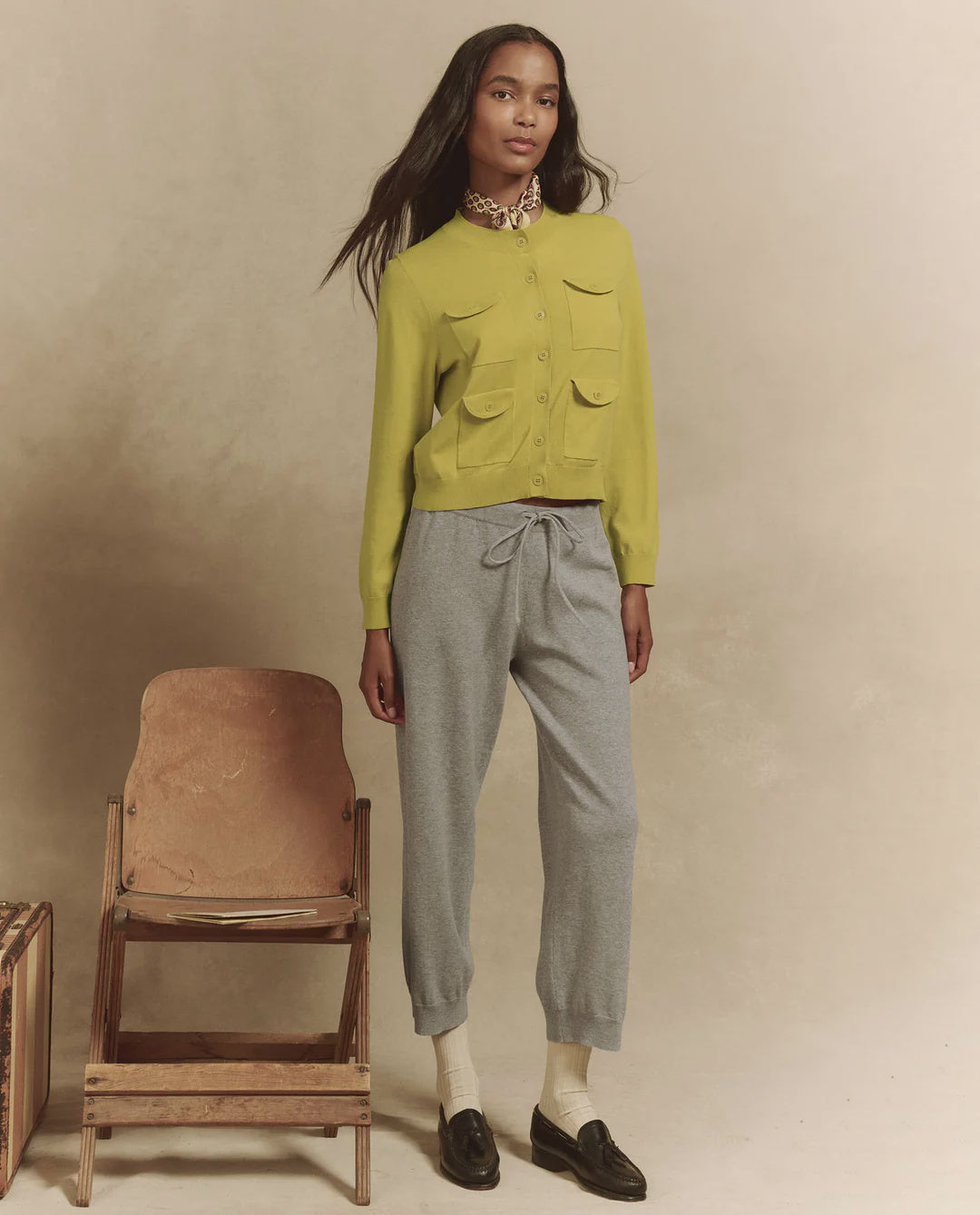 A person stands beside a vintage wooden chair wearing The Great Inc.'s yellow-green Patch Pocket Cardigan, grey drawstring pants, white socks, and black loafers against a neutral beige background.