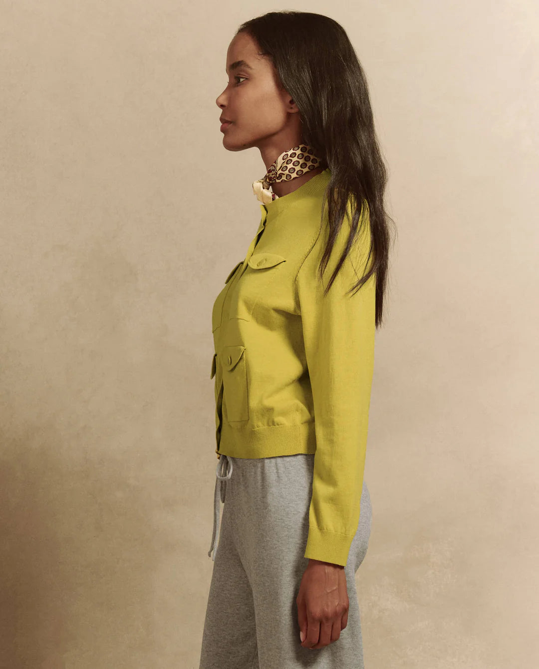 A woman in profile wears The Great Inc.'s slim-fit Patch Pocket Cardigan with utility-inspired pockets in yellow, paired with a patterned scarf and gray pants against a neutral background. Her long hair cascades naturally over her shoulders.