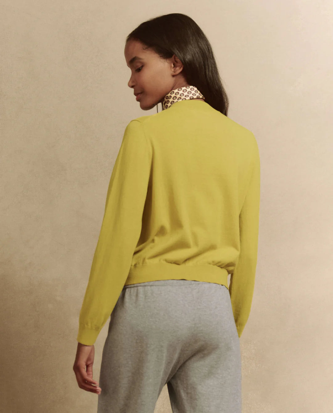 A person with long dark hair wears The Patch Pocket Cardigan by The Great Inc., a slim fit mustard yellow sweater crafted from 100% cotton, featuring a white and brown patterned collar. They are paired with gray pants and stand against a beige background, facing sideways.