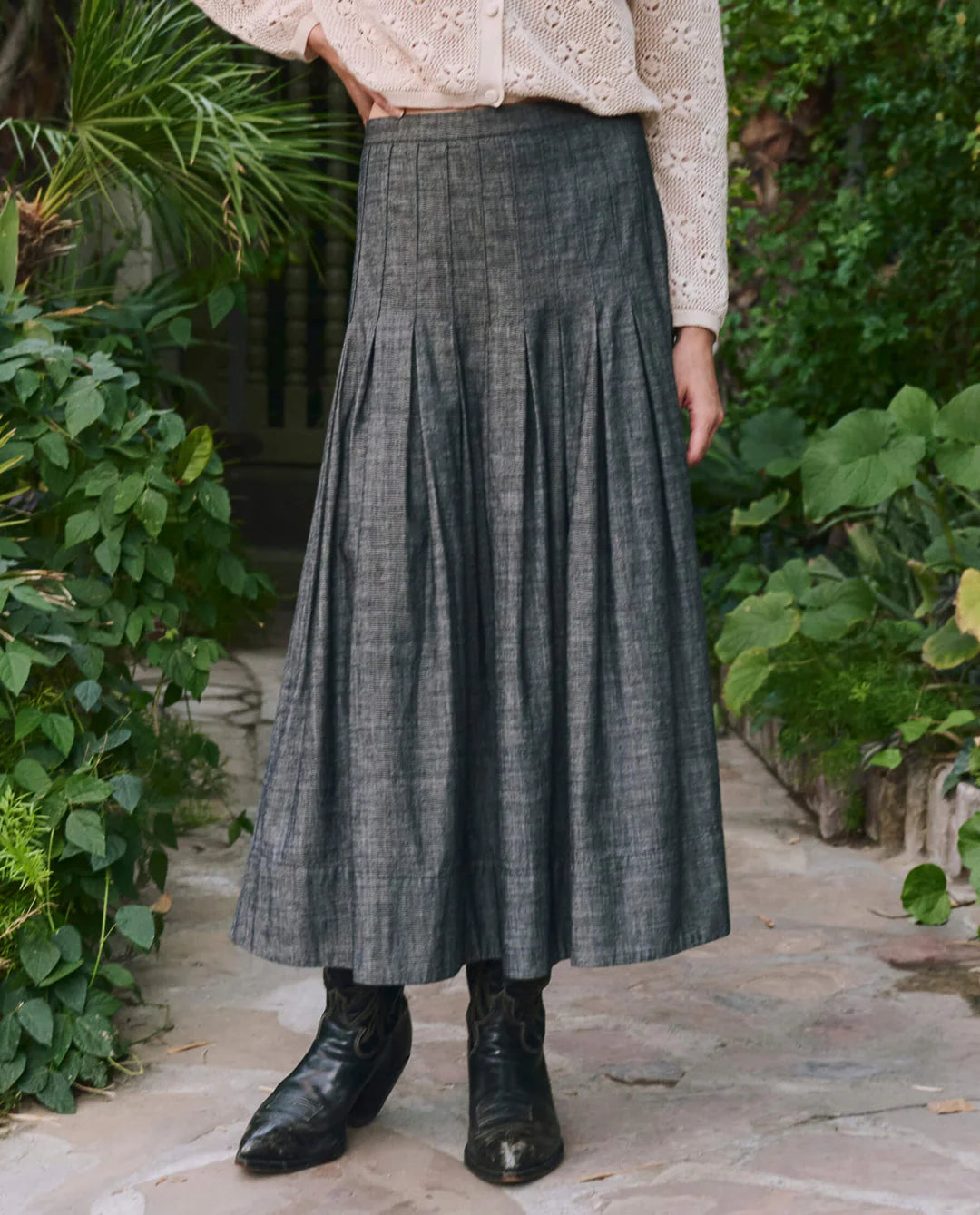 A person wearing The Great Inc.'s The Rhythm Skirt, a high-waisted gray midi, paired with a cream lace blouse and black boots, stands on a stone path among green plants.