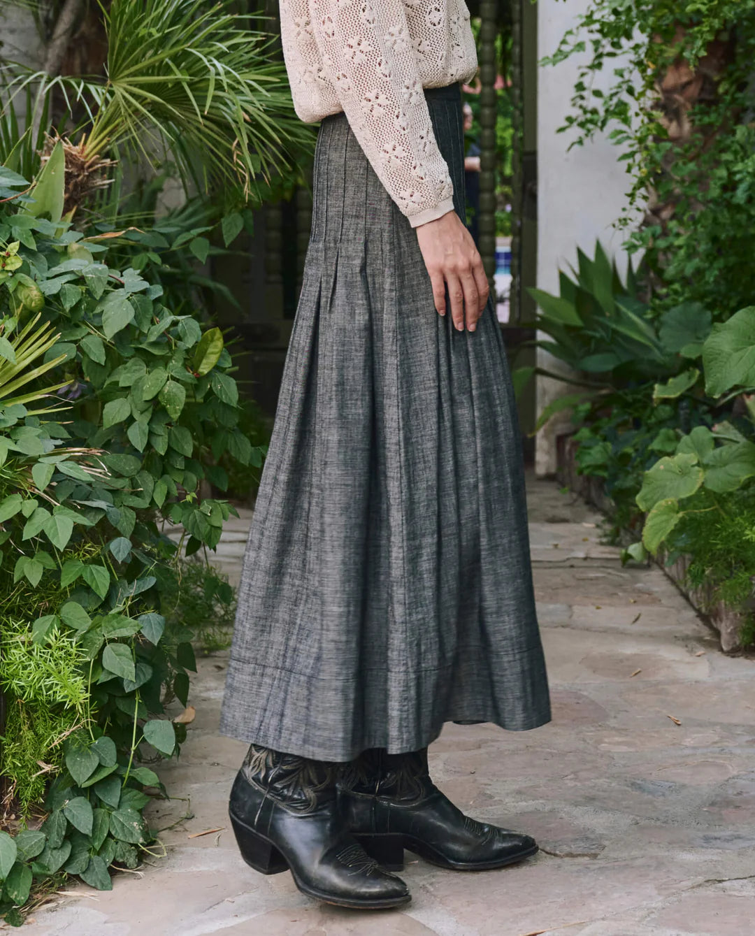A person wearing The Rhythm Skirt by The Great Inc., a textured gray baby wale cord midi, with a cream lace long-sleeve top and black leather cowboy boots stands on a stone path amid lush green plants.