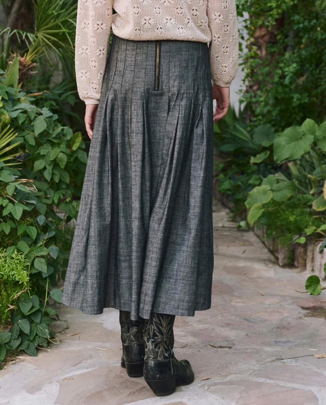 A person stands outdoors among greenery, wearing The Rhythm Skirt by The Great Inc., paired with black floral boots and a cream lace-texture long-sleeve top.