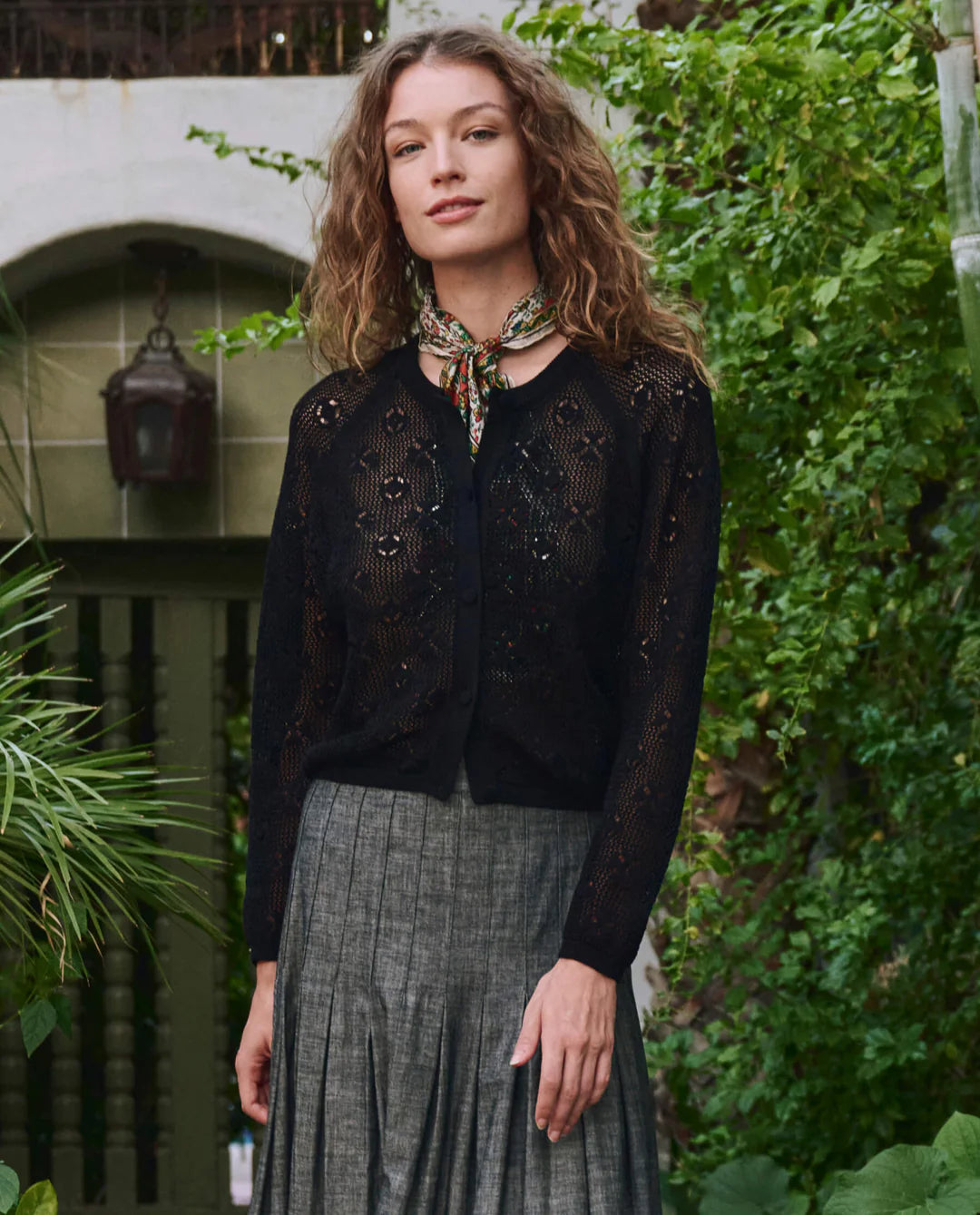 A woman with wavy hair wears The Great Inc.'s The Crochet Cardigan, styled with a gray pleated skirt and patterned scarf. She stands in a lush garden, green plants and a stone archway behind her.