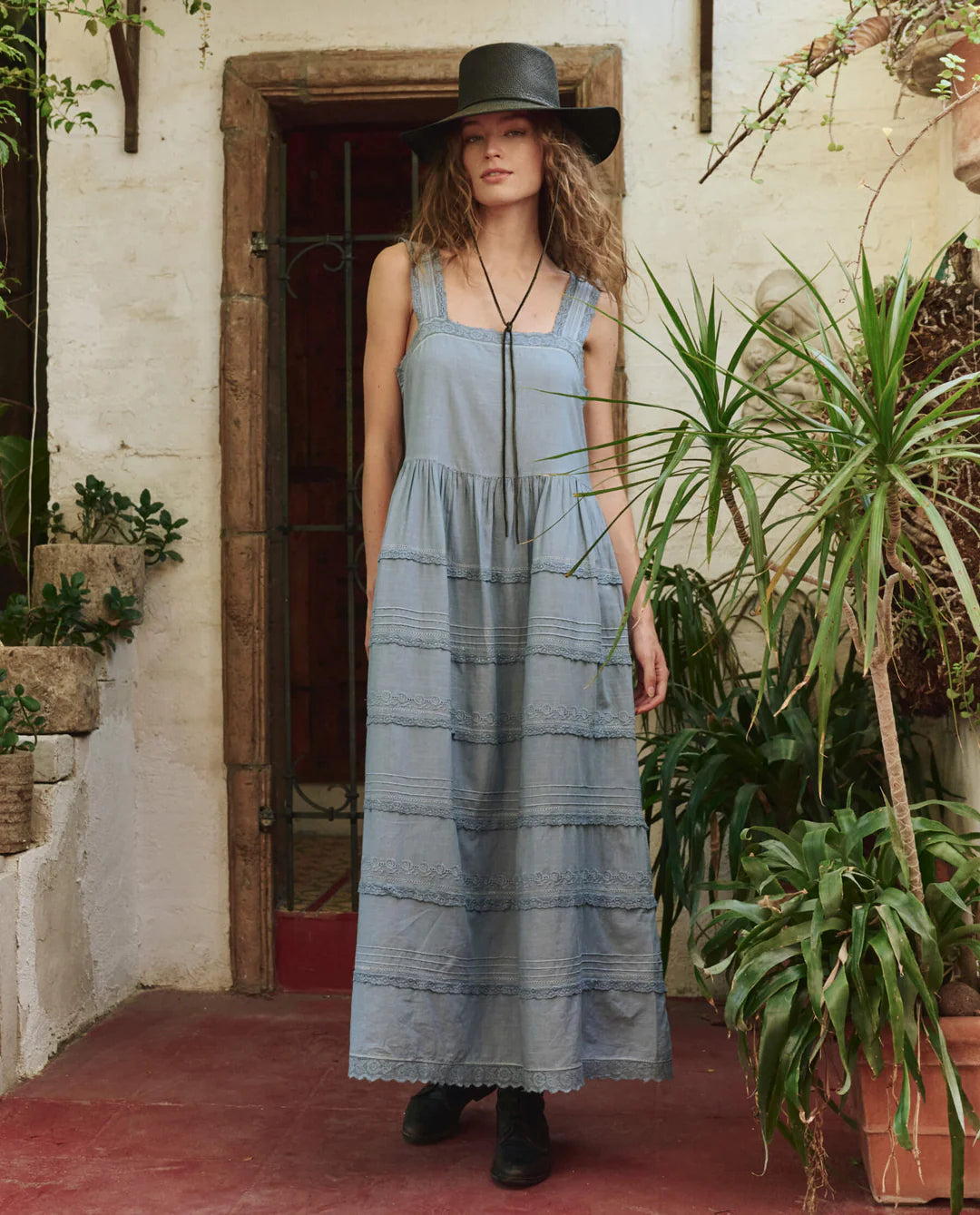 A woman wears The Reverie Dress by The Great Inc.—a long, sleeveless blue cotton dress—paired with a wide-brimmed black hat. She stands on a red floor among green plants, beside a white wall and wooden door. Dry clean recommended.