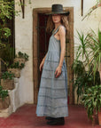 A woman stands sideways on a red-tiled floor among green plants, wearing The Reverie Dress by The Great Inc.—a long, sleeveless blue cotton dress—paired with a black hat, in front of a rustic door and white wall. Dry clean recommended.