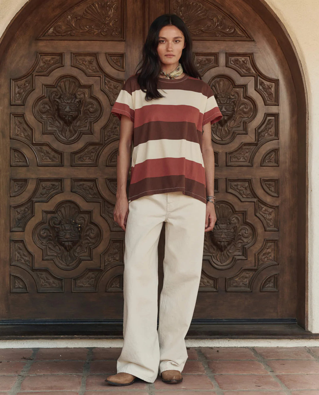 A woman stands before an ornate wooden door in The Boxy Crew 26 by The Great Inc., paired with cream wide-leg pants and brown shoes—effortlessly channeling a handcrafted Los Angeles aesthetic.