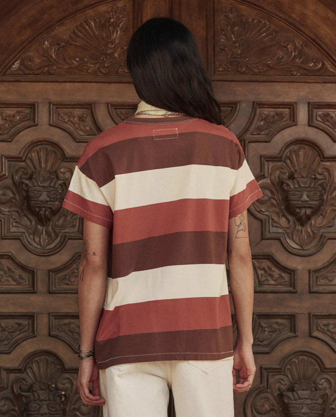 A person with long dark hair, wearing The Boxy Crew 26 by The Great Inc.—a short-sleeved vintage tee in brown, rust, and cream stripes—and light-colored pants stands facing an ornate carved wooden door.
