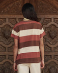 A person with long dark hair, wearing The Boxy Crew 26 by The Great Inc.—a short-sleeved vintage tee in brown, rust, and cream stripes—and light-colored pants stands facing an ornate carved wooden door.