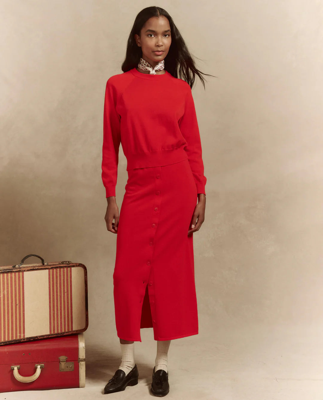 A person wearing The Crew Neck Sweater from The Great Inc., in red high-twist yarn, paired with a matching buttoned skirt, poses confidently next to vintage suitcases. A neutral background highlights their classic, stylish look.