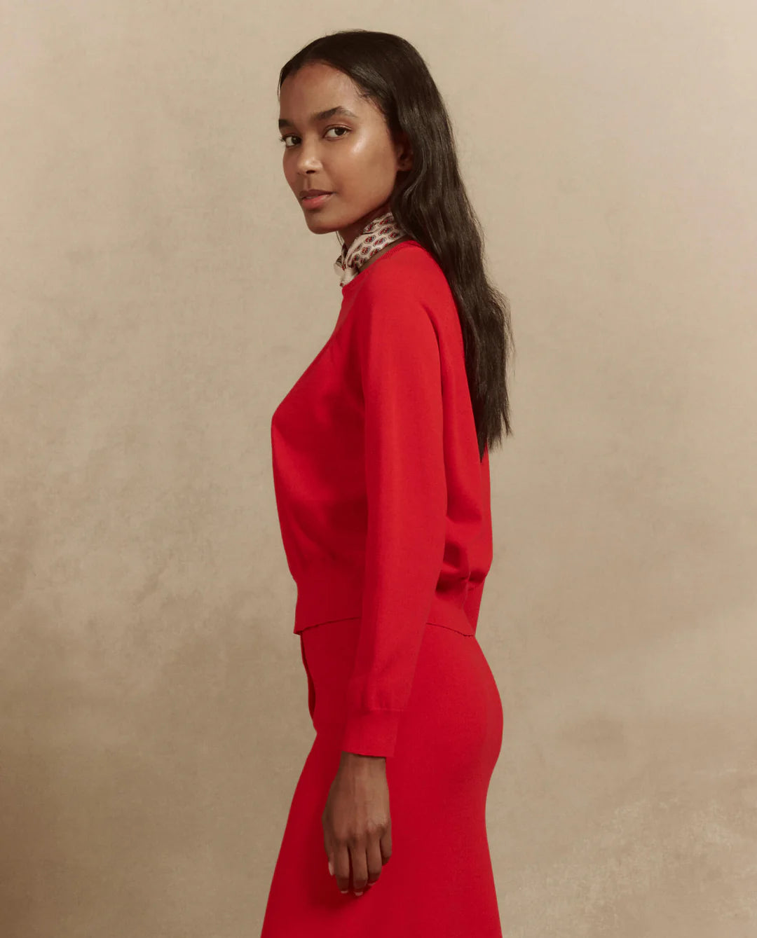 A woman with long hair, dressed in The Great Inc.'s red "The Crew Neck Sweater," stands against a neutral background. She gazes calmly at the camera, highlighting the sweater's versatility and fine gauge yarn design.