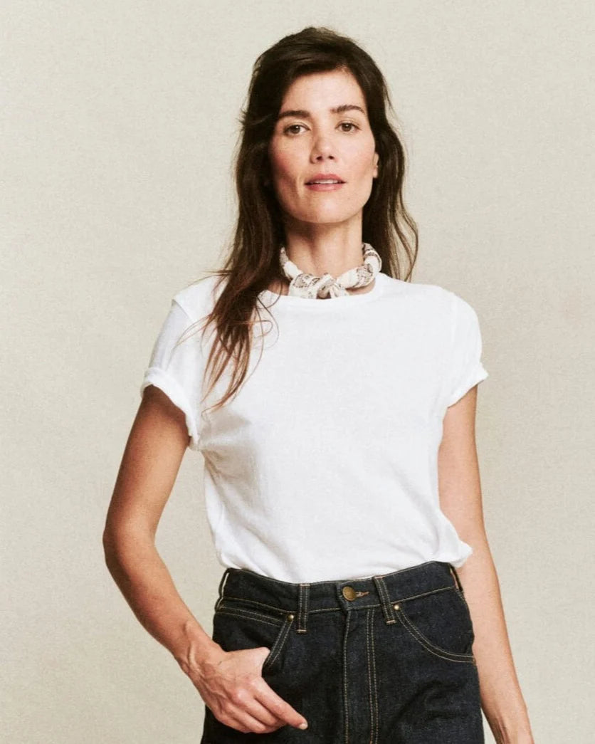 A woman with long brown hair models The Little Tee by The Great Inc., paired with dark high-waisted jeans and a chunky white necklace, standing against a plain beige background.