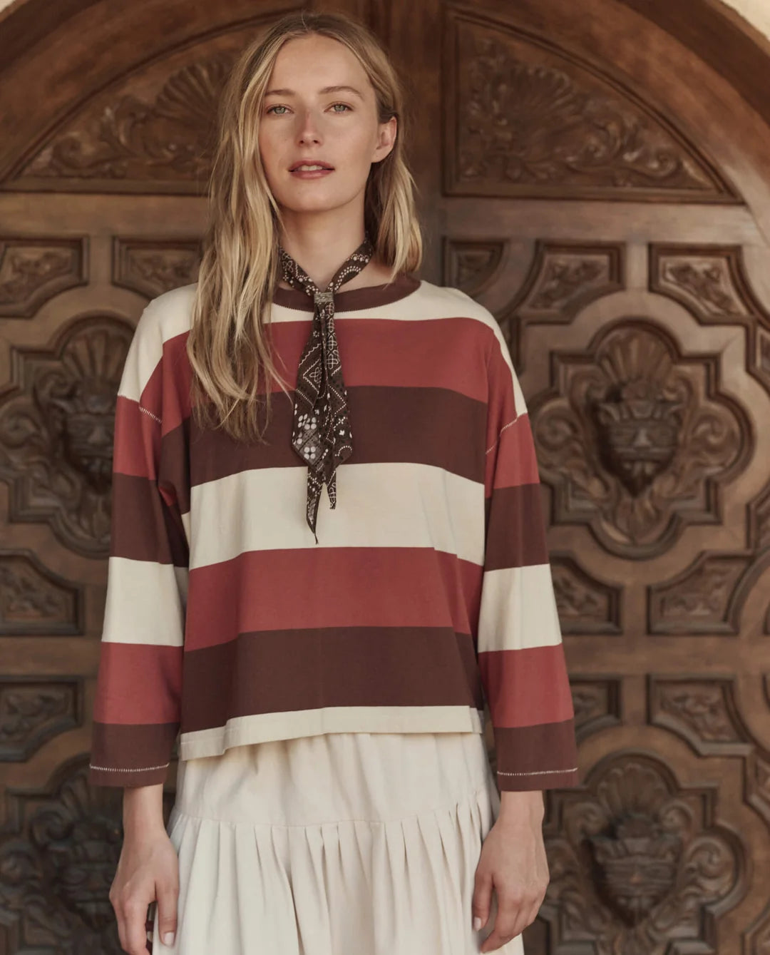 The Great Inc. Field Tee in striped pattern, styled with a bandana scarf, by an ornate wooden doorway.