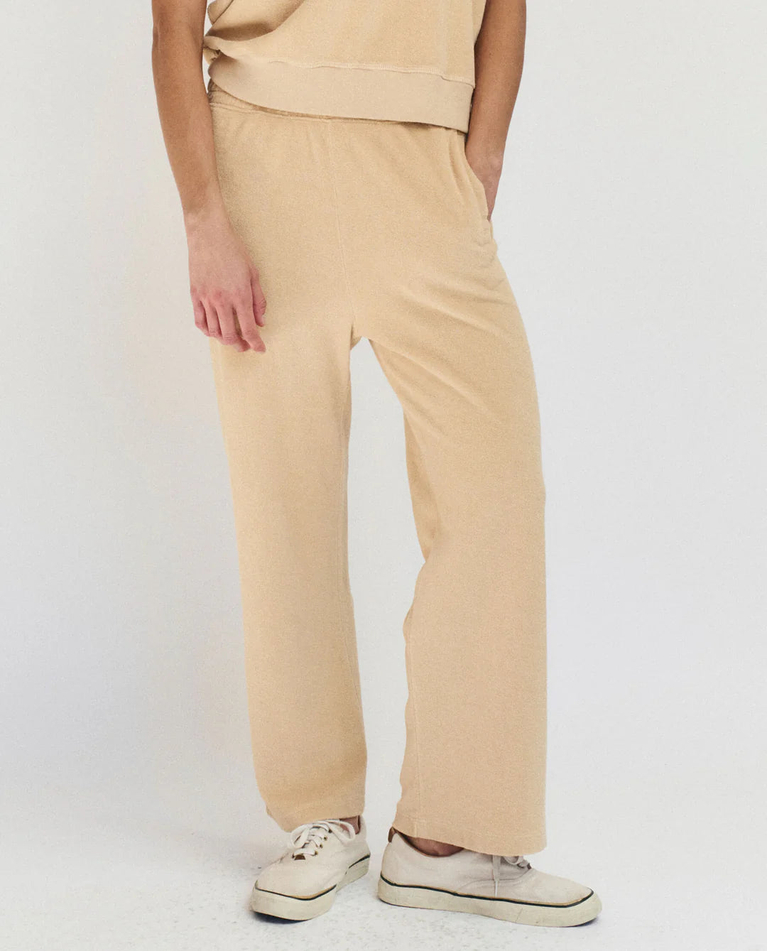 A person stands against a plain background, wearing The Micro Terry Sweatpant by The Great Inc. in loose beige cotton, paired with a matching beige top and white sneakers; image is cropped from the shoulders down.