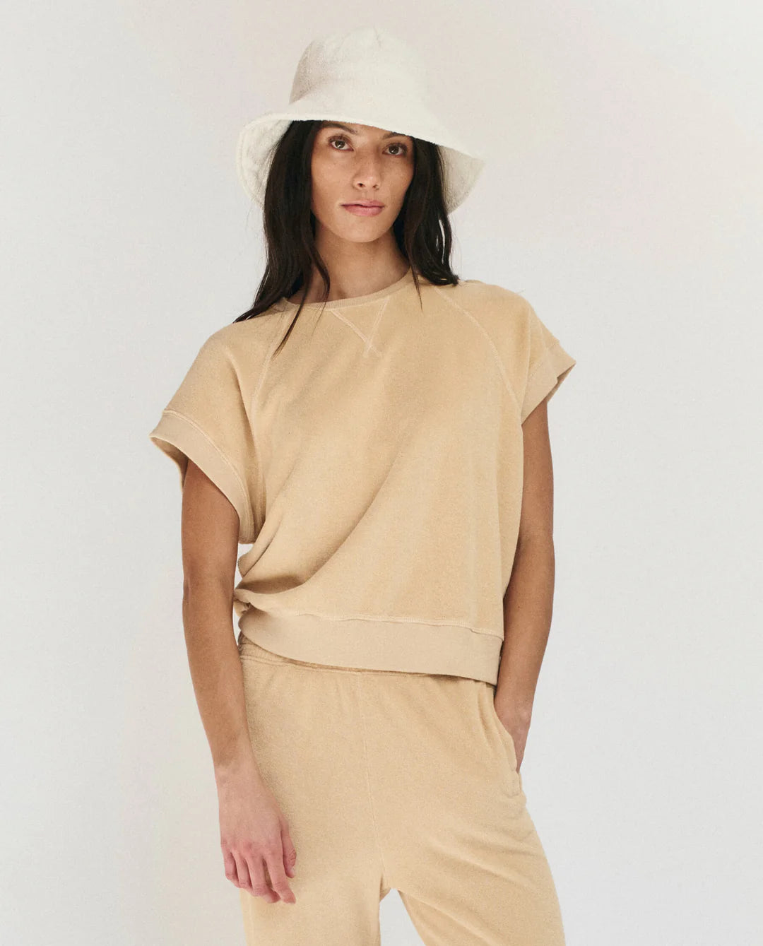 A woman with long dark hair wears The Micro Terry Wedge SS by The Great Inc., a beige short-sleeve top with matching pants, styled with vintage knits and a cream bucket hat, standing against a plain white background.