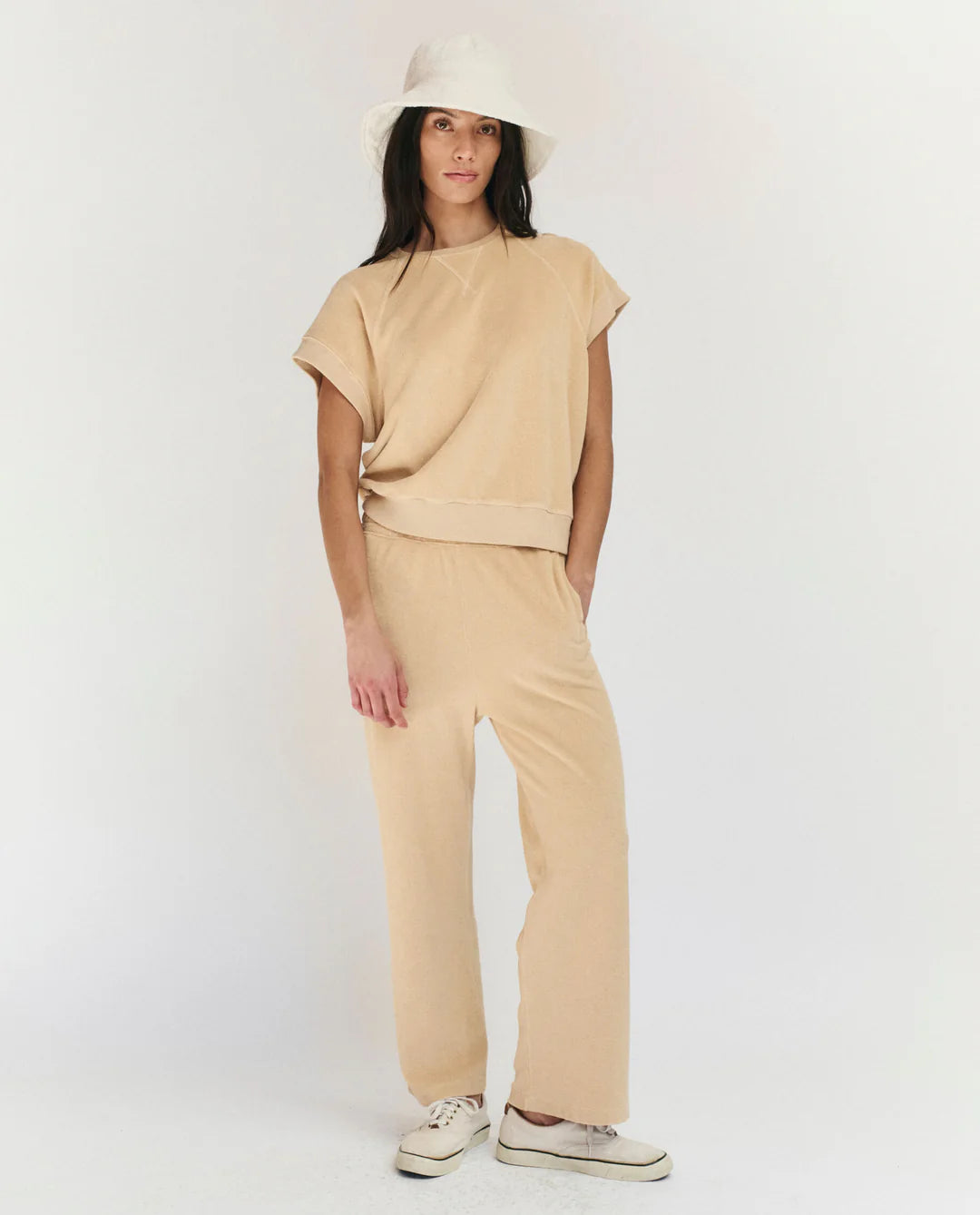 A woman wearing The Great Inc. Micro Terry Sweatpant in beige with a matching short-sleeve top, white bucket hat, and white sneakers stands against a plain white background.