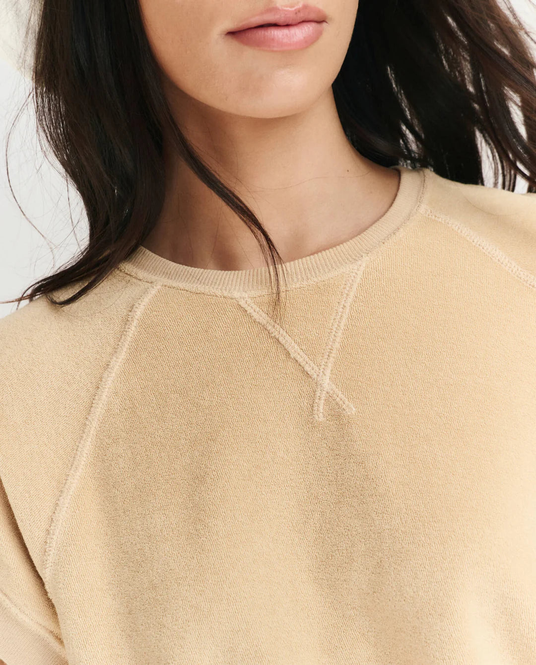 A woman with long dark hair wears The Micro Terry Wedge SS by The Great Inc., a beige sweatshirt with visible seam details. The photo captures only her lower face and upper torso, emphasizing its subtle vintage-inspired style.