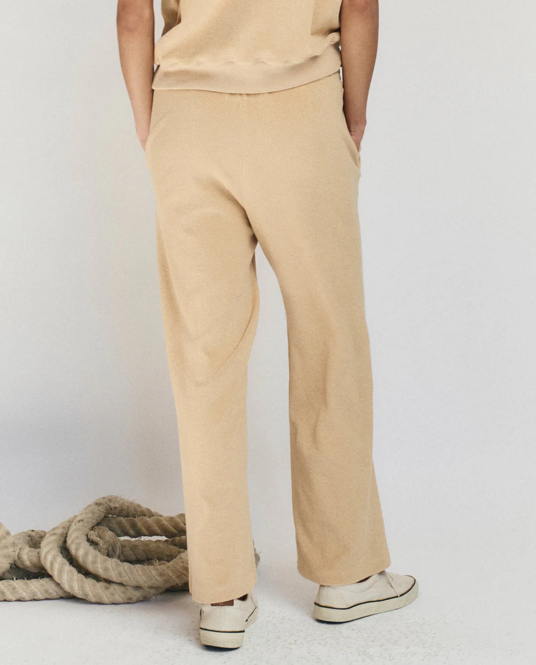 A person stands facing away, wearing The Micro Terry Sweatpant by The Great Inc. in beige with a matching top and white sneakers, hands in pockets. A thick coiled rope lies on the floor against a plain background.