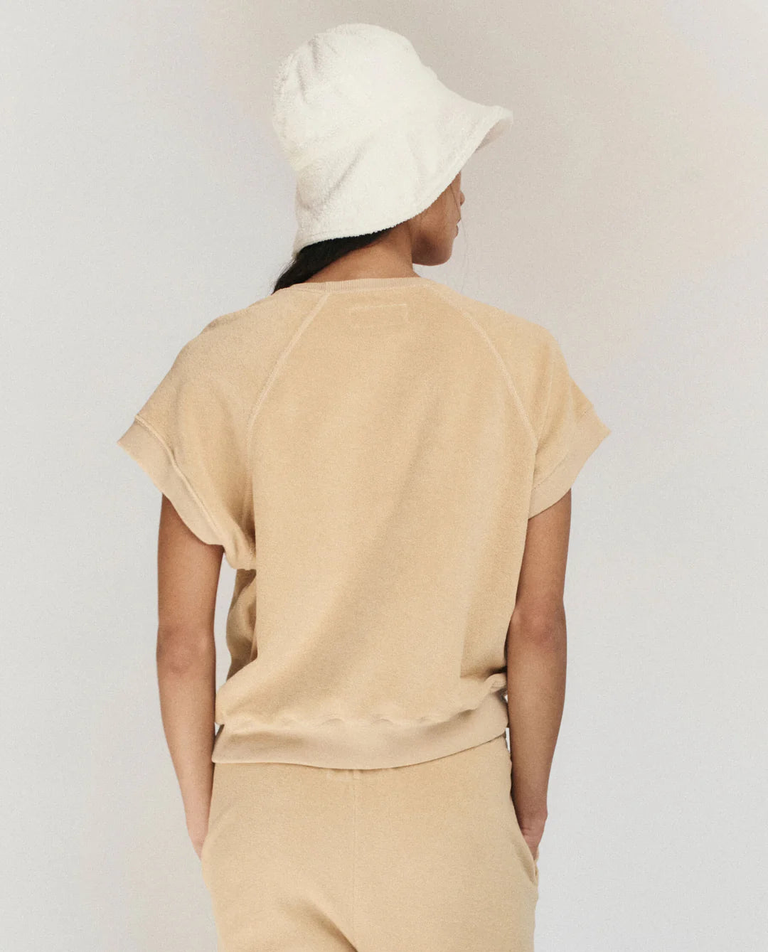 A person faces away from the camera, wearing The Great Inc.'s The Micro Terry Wedge SS in beige with matching pants and a white bucket hat, set against a plain light background.