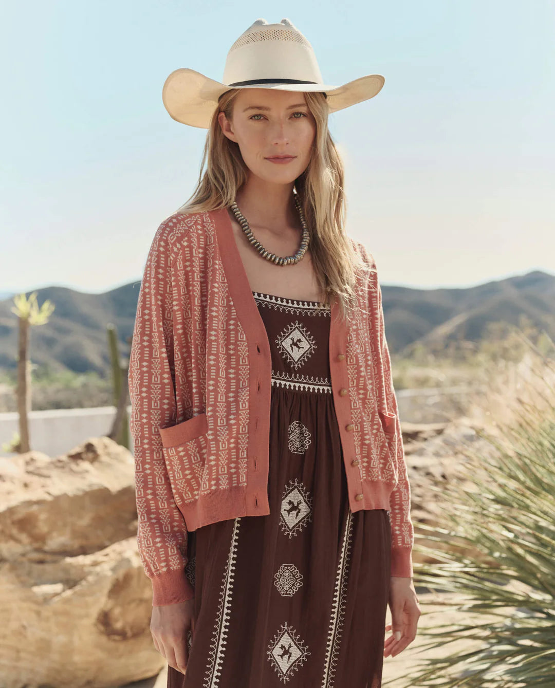 A woman wears The Great Inc.'s The Varsity Cardigan over a patterned brown dress and white cowboy hat, standing outdoors among mountains and desert plants, looking at the camera.