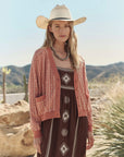 A woman wears The Great Inc.'s The Varsity Cardigan over a patterned brown dress and white cowboy hat, standing outdoors among mountains and desert plants, looking at the camera.