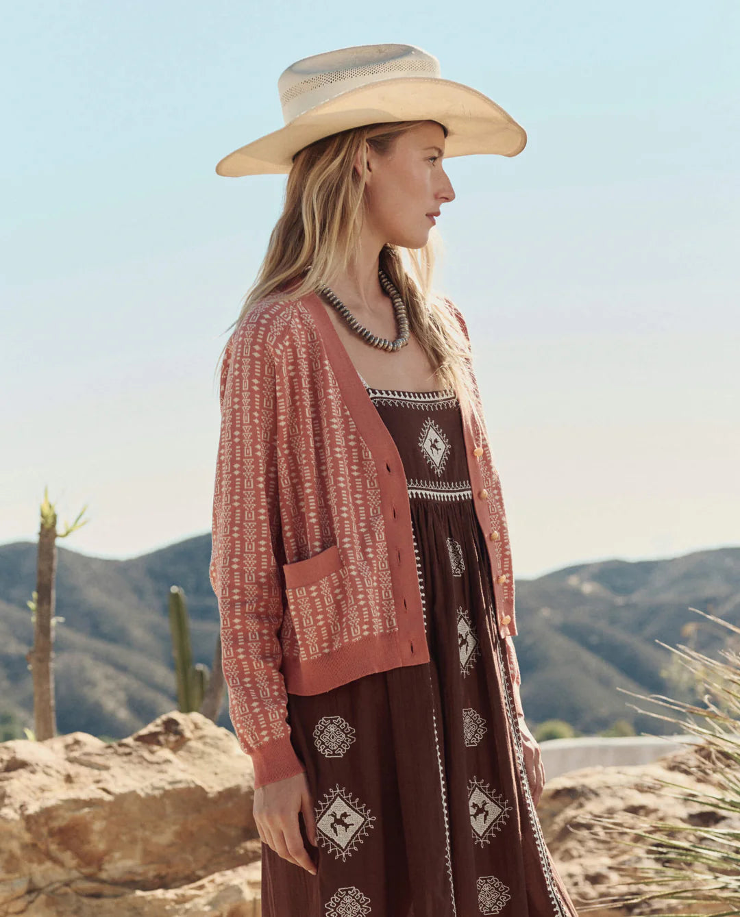 A woman in The Great Inc.'s The Varsity Cardigan, styled with a cowboy hat and brown dress, stands outdoors in a desert landscape with mountains and cacti behind her.