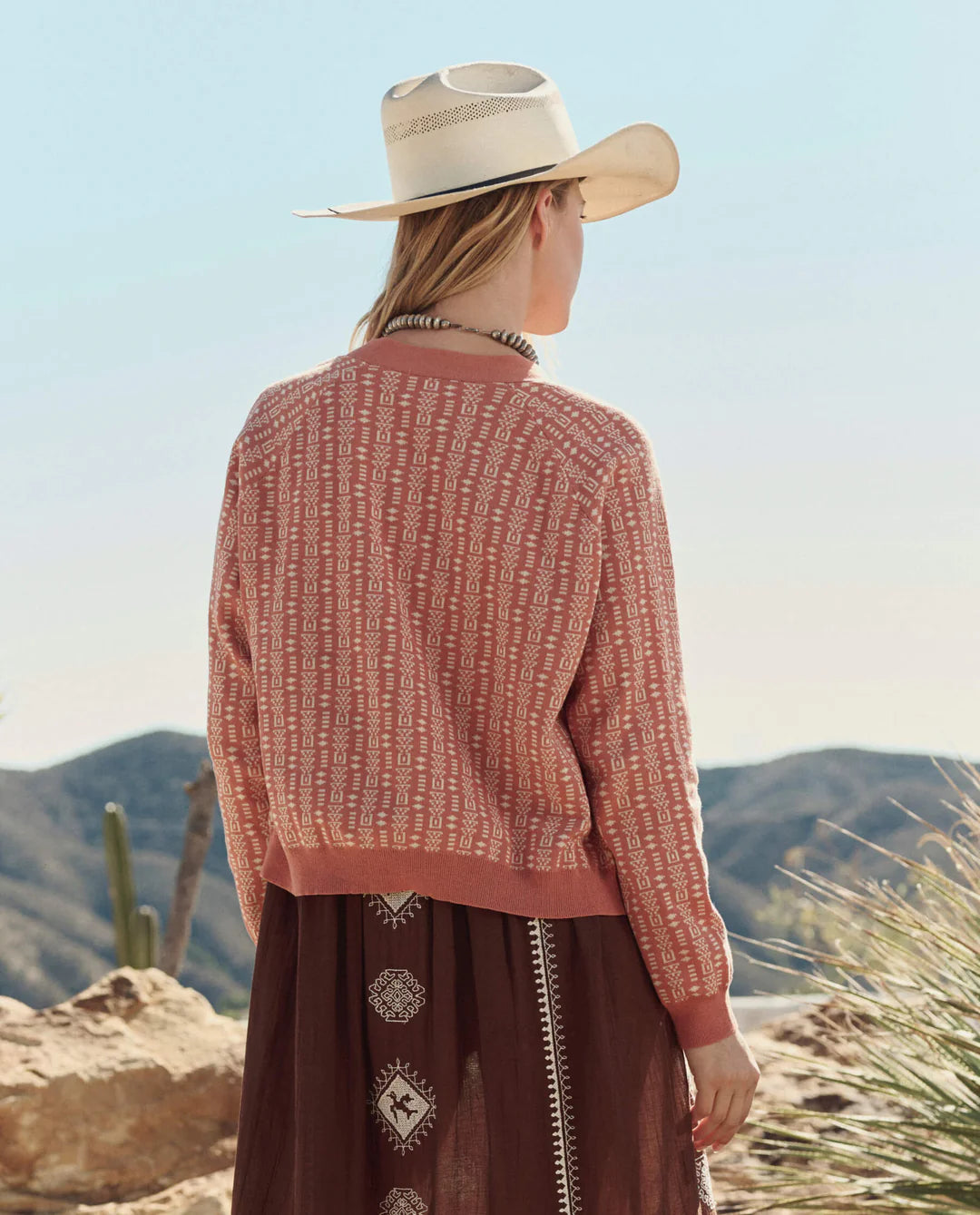 A woman in The Great Inc.'s The Varsity Cardigan stands outdoors with her back to the camera, facing desert hills and cacti under a clear sky.